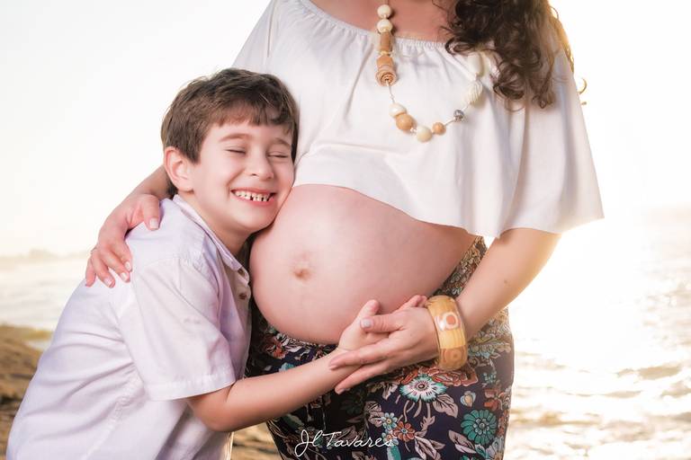 Menino sorrindo abraçando a barriga da mãe grávida durante o ensaio gestante na praia.