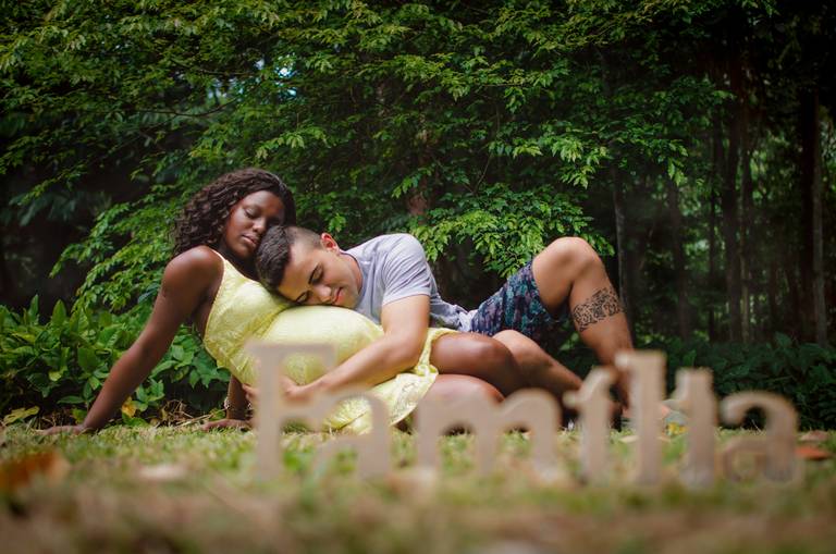 Casal sentado na grama do Jardim Botânico do Rio de Janeiro, marido abraçando a barriga da esposa no detalhe uma placa escrito familia em primeiro plano desfocada!