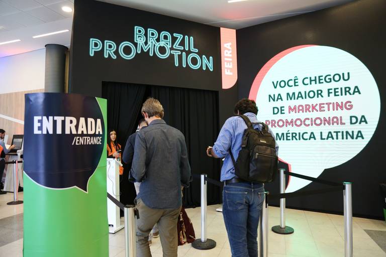 Entrada principal de uma feira de negócios, com sinalização e fluxo de pessoas chegando ao local.