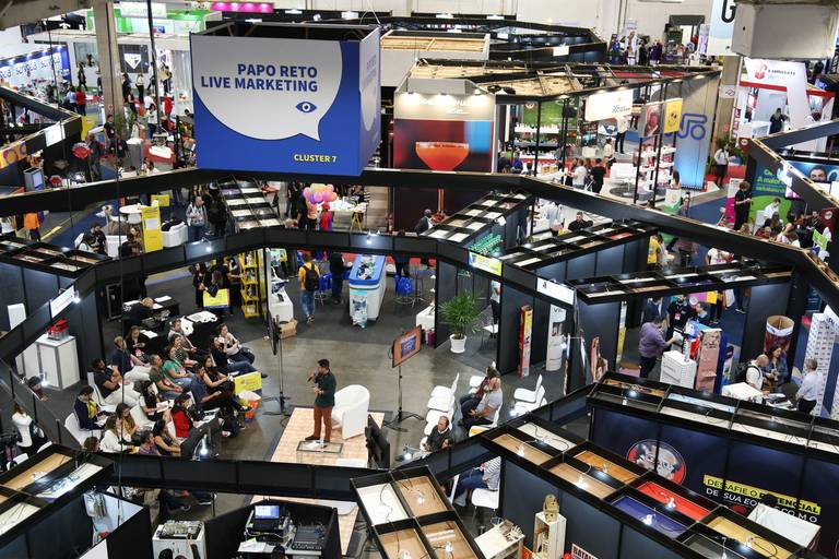 Vista aérea do saguão de uma feira, com grande movimentação de pessoas e estandes distribuídos.