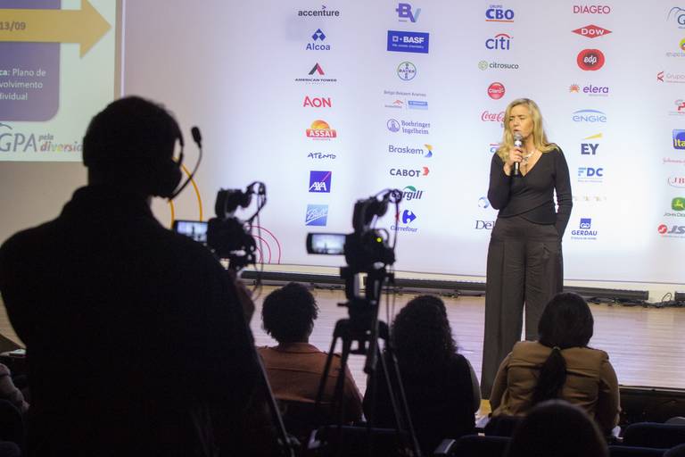 Palestrante falando no palco, enquanto uma câmera profissional o filma, indicando a gravação do evento.