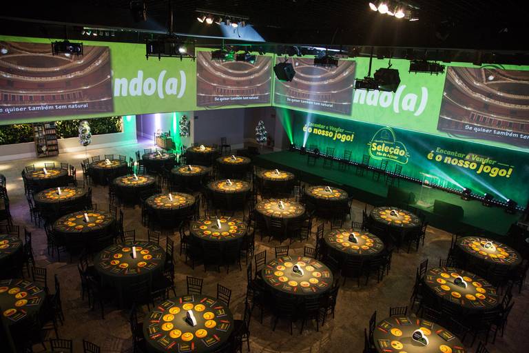 Vista aérea de cima das mesas e do palco de um evento, antes do início, mostrando a organização e o espaço pronto para receber o público.