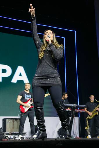 Cantora em destaque no palco, com microfone em mãos, interpretando uma canção em um evento.