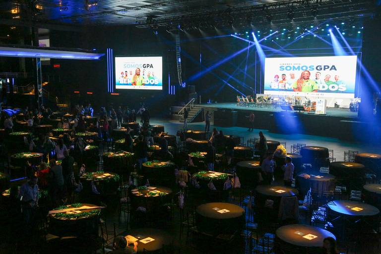 Salão de evento decorado com mesas dispostas e um palco visível ao fundo, pronto para apresentações.