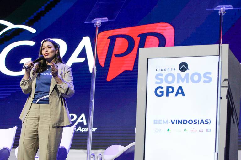Pessoa falando no palco, com foco na sua comunicação e no ambiente de apresentação do evento.