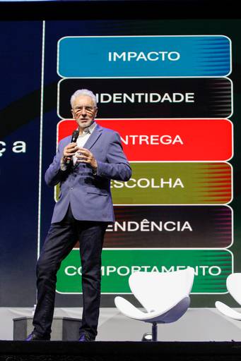 Palestrante em primeiro plano, com iluminação ou enquadramento que o coloca em grande destaque no palco.