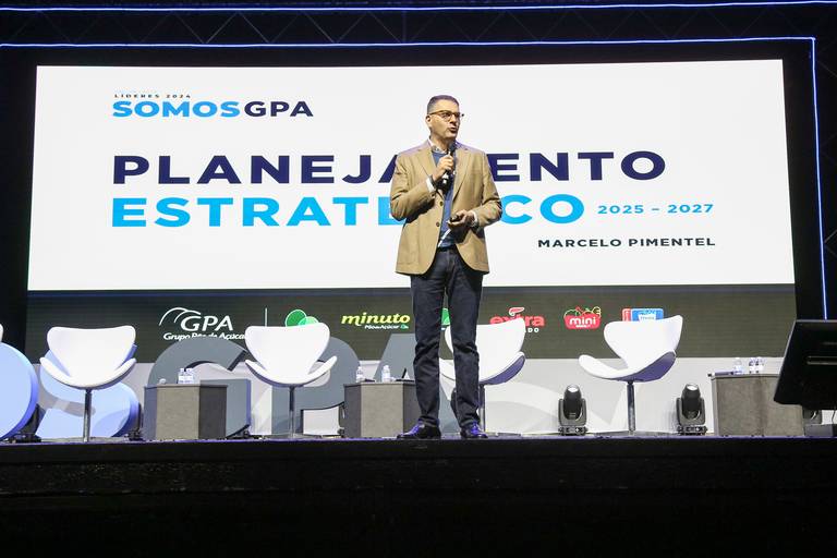 Pessoa falando no palco, com microfone, direcionando-se à audiência em um evento.