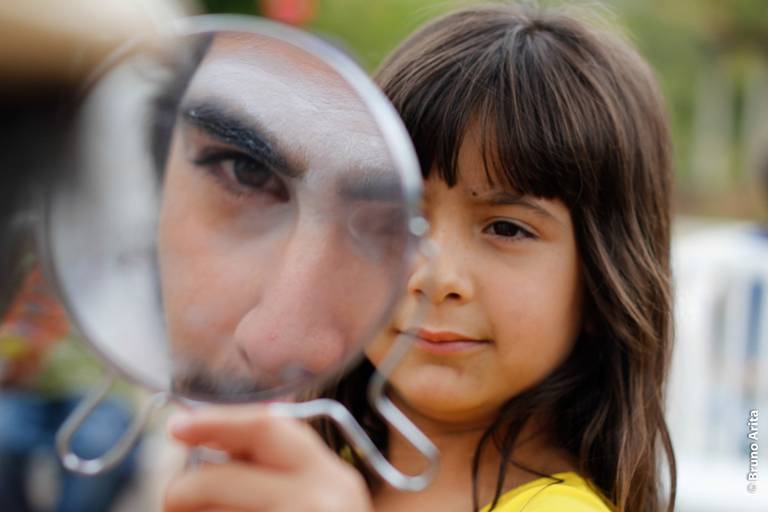 Menina segurando um espelho para um palhaço, que está se maquiando, em um momento divertido e de bastidores.