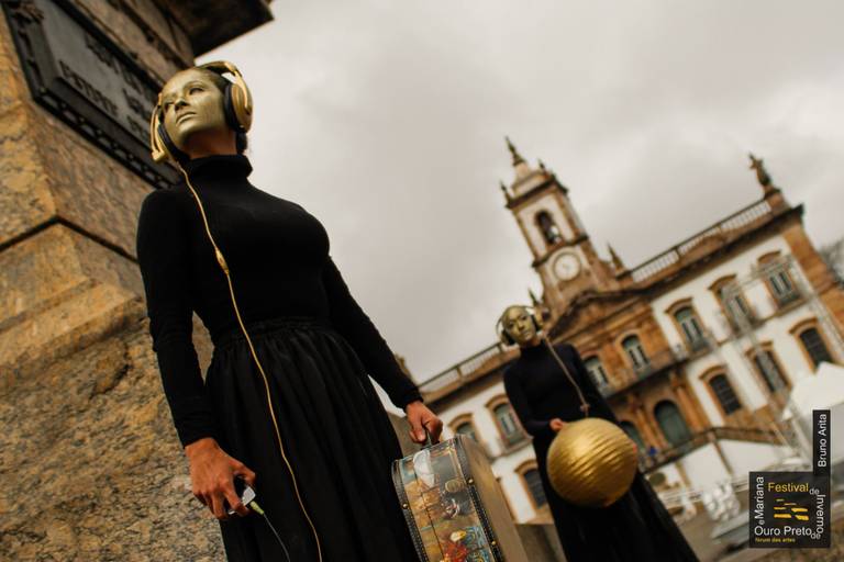 Duas artistas performáticas no meio de uma cidade, com figurinos e adereços que chamam a atenção.