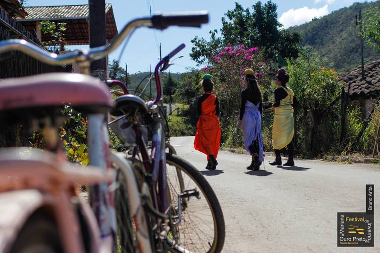 Três artistas andando em uma cidade, possivelmente em um cortejo ou intervenção urbana, com figurinos ou adereços.