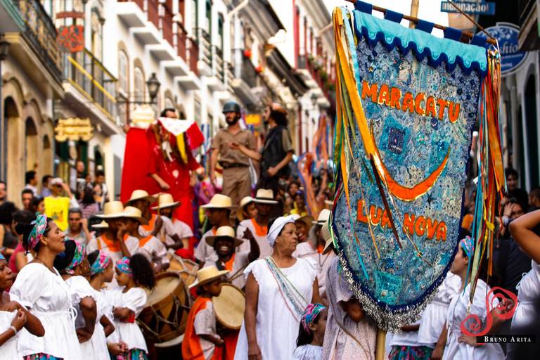 Cortejo cultural ou festivo nas ruas, com pessoas caminhando, possivelmente com música e dança.