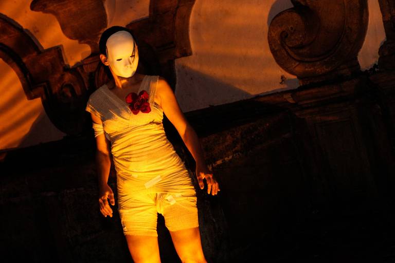 Artista usando máscara iluminada por uma luz quente, criando um efeito dramático e misterioso em uma apresentação.