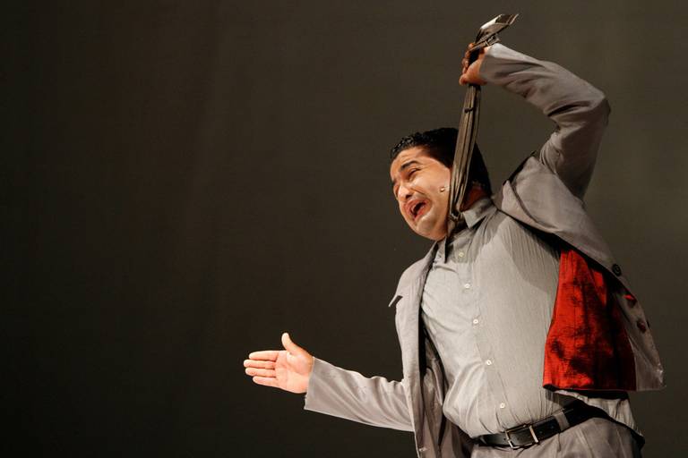 Artista performático segurando a própria gravata, em um gesto teatral que sugere um puxão ou movimento dramático.