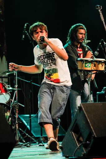 Cantor de reggae se apresentando no palco, com microfone e instrumentos ao fundo, transmitindo a energia do show.
