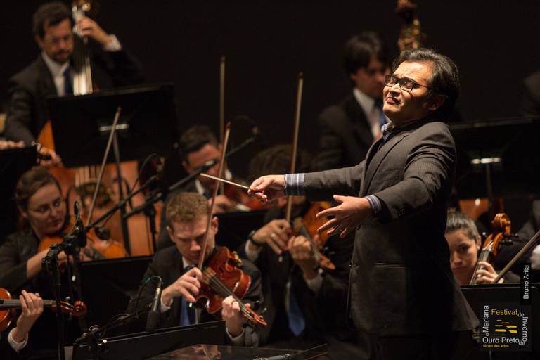 Maestro regendo uma orquestra ou grupo de músicos, com os instrumentistas em segundo plano, em um concerto.