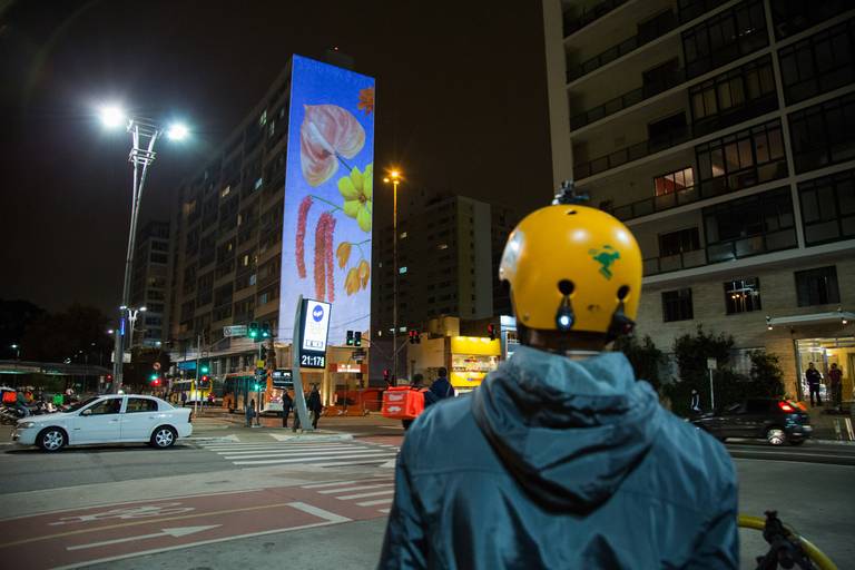 Ciclista com capacete observando uma projeção artística em um prédio, em um momento de contemplação urbana.