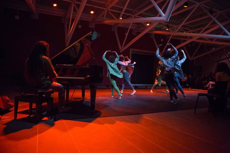 Dançarinos performando ao fundo, enquanto um pianista os observa, criando uma cena de interação artística.