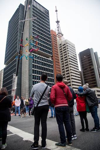 Pessoas na rua observando atentamente um espetáculo de artistas suspensos, com olhares curiosos e admirados.