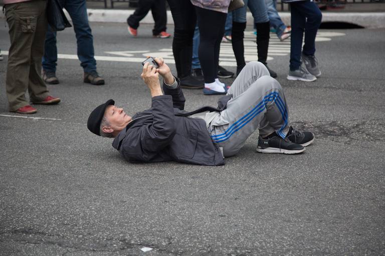 Homem da plateia deitado no chão para fotografar um espetáculo cultural, buscando um ângulo único para o registro.