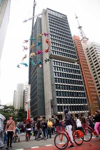 Artistas suspensos em performance aérea sobre uma avenida, como parte de um espetáculo cultural ao ar livre, observados pelo público.