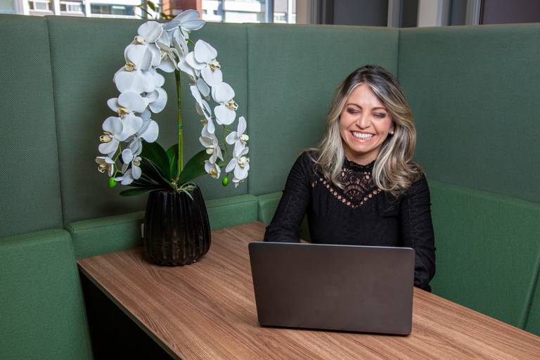 Mulher sorrindo, engajada no trabalho com seu notebook na estação, com orquídea na mesa, em retrato no ambiente de trabalho. (Retrato Corporativo, São Paulo, São Paulo, Fotógrafo Bruno Arita)