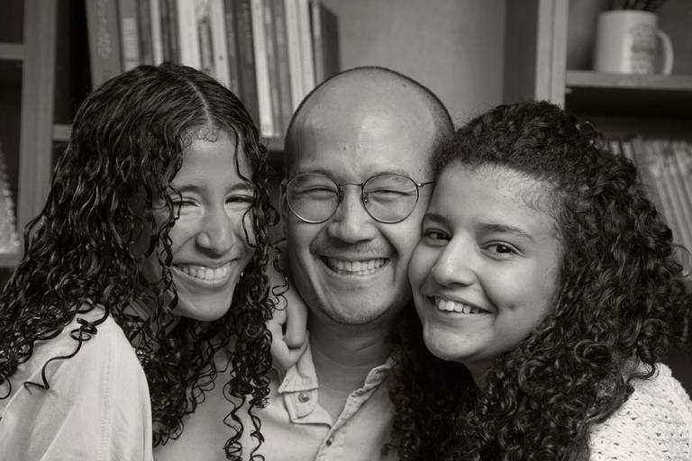 Pai sorrindo com suas duas filhas, em uma foto que transmite alegria e conexão familiar.