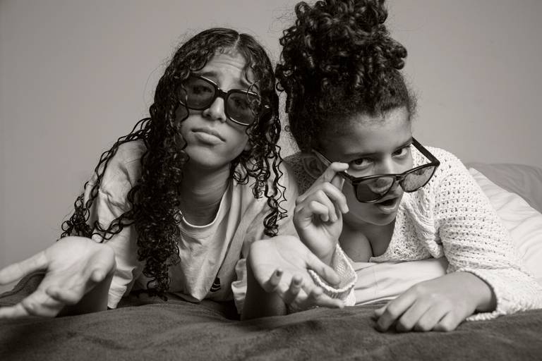 Duas filhas usando óculos, posando de forma divertida para uma foto na cama, em um ensaio familiar.