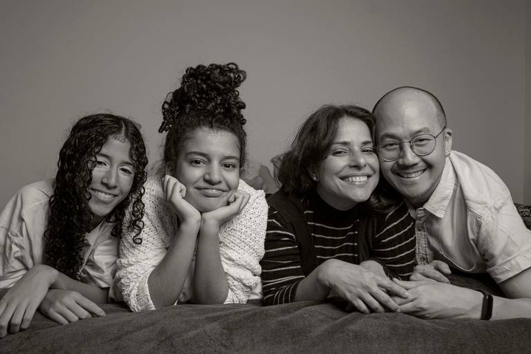 Toda a família posando para uma foto na cama, em um cenário aconchegante e descontraído do ensaio.
