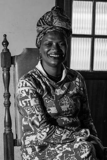 Retrato P&B da modelo sorrindo e usando turbante, evocando a tradição e resistência da mulher negra em Ouro Preto. Foto por Bruno Arita