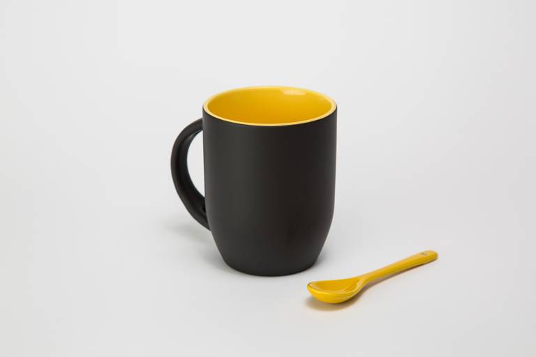 Fotografia still de caneca para personalização na cor preta na parte externa e amarelana parte interna com colher amarela acompanhando o kit. Foto por Bruno Arita.