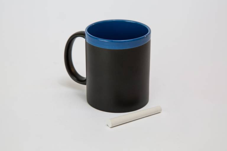 Fotografia still de caneca para personalização na cor preta na parte externa e azul na parte interna com giz branco ao lado para escrever na caneca. Foto por Bruno Arita.