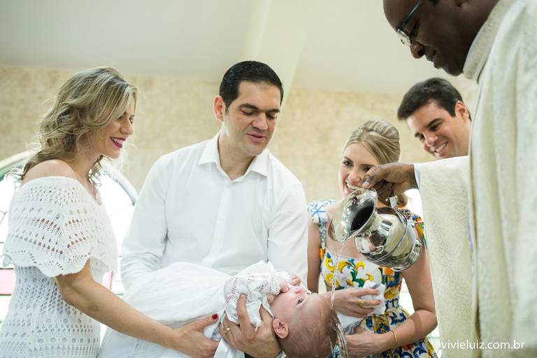 padre batizando o bebê, que está sendo segurado pelos padrinhos, com água 