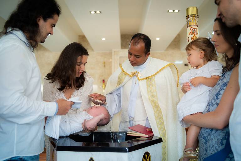 padre batizando o bebê, que está no colo da madrinha, com água