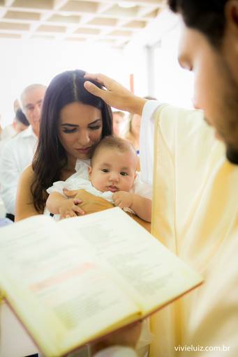 padre dando benção no bebê que stá sendo segurado pela madrinha, no batizado