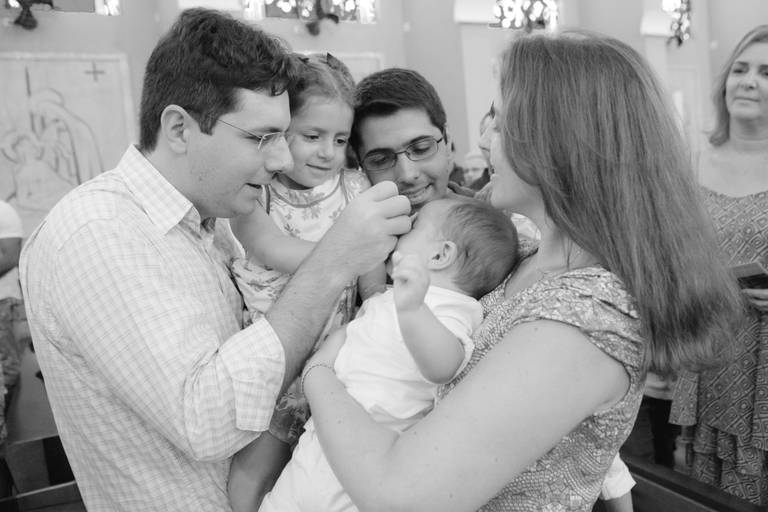 pai fazendo sinal da cruz no filho com irmã mais velha no colo durante batizado realizado na Paróquia São João Bosco, Americana, São Paulo