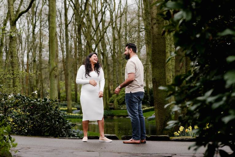 Candid maternity photo of a couple laughing together during a Keukenhof photoshoot, set among tall trees and spring flowers. Ideal for travelers visiting the Netherlands who book a babymoon session near Amsterdam