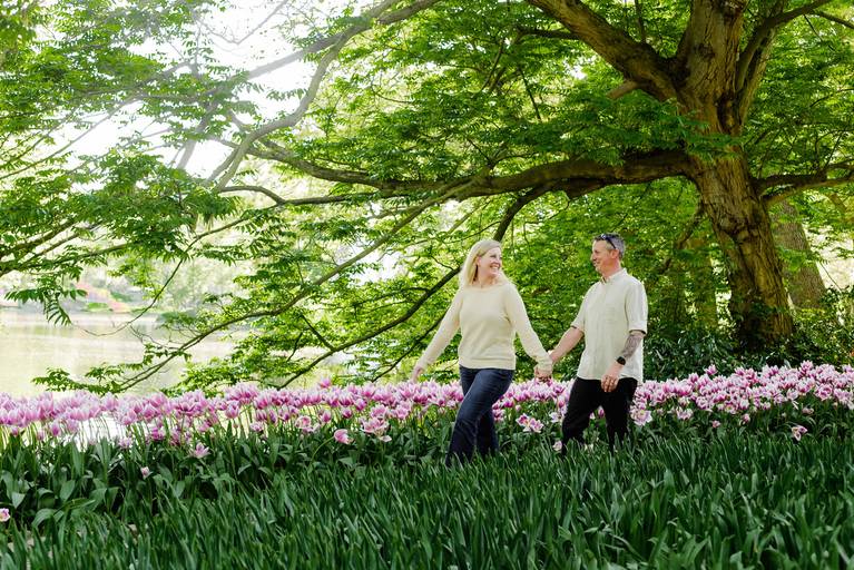 Couple walking hand in hand under the shade of tall green trees, with rows of pink tulips beside them. A romantic photoshoot experience for travelers exploring the Netherlands in spring.