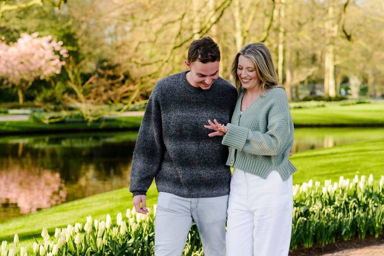Couple laughing together by a calm pond in Keukenhof Gardens as she shows her engagement ring. Captured moments after a surprise proposal in a quiet area chosen with the help of an experienced proposal photographer.