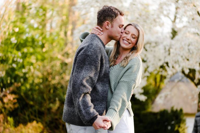 Couple smiling as he kisses her cheek after a surprise proposal in Keukenhof Gardens. Soft spring light and blooming trees frame the moment. Captured by an experienced Keukenhof proposal photographer who guides clients to quiet spots