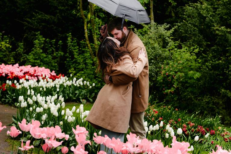 Couple holding umbrellas during a rainy proposal session in Keukenhof, standing between bright yellow tulips. The moment is intimate and relaxed, photographed in a sheltered path to avoid crowds