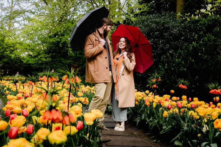 Couple kissing under a shared umbrella between rows of pink and white tulips at Keukenhof. A private proposal moment captured in a quiet part of the garden