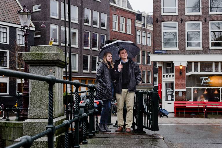 Couple standing under an umbrella on a rainy canal bridge in Amsterdam, perfect for travelers planning a cozy and romantic proposal in the city.
