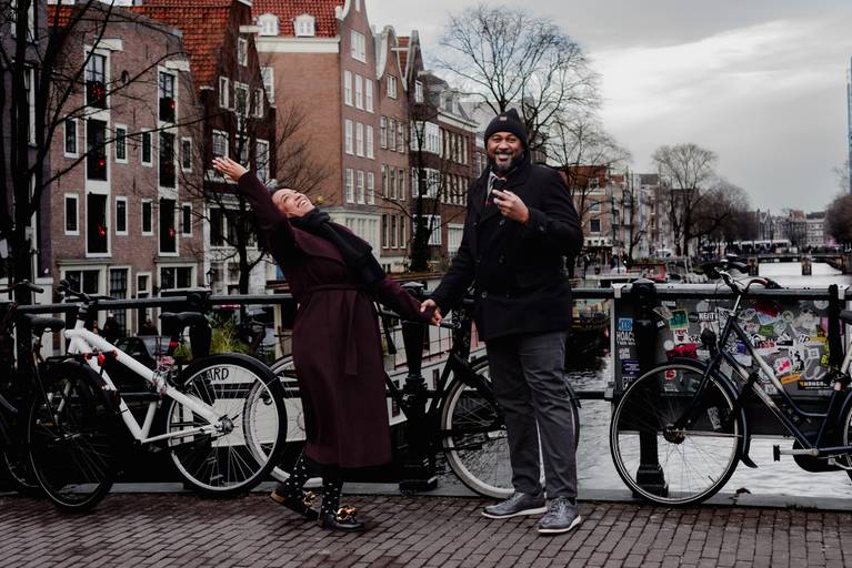 Joyful pair celebrating on an Amsterdam bridge with bikes behind them, perfect for travelers looking for fun and natural couple photography in the city.