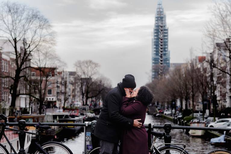 Couple kissing on an Amsterdam bridge with canal views and bikes, perfect for travelers planning a surprise engagement or booking a local photographer.