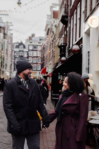 Couple holding hands and walking through a cozy Amsterdam street with string lights overhead, ideal for travelers seeking engagement photo ideas or a local photographer.