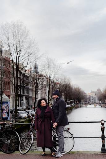 Couple smiling on a winter bridge in Amsterdam, standing by bikes along the canal. Perfect moment for travelers looking for a romantic photoshoot or inspiration for a proposal in the city.