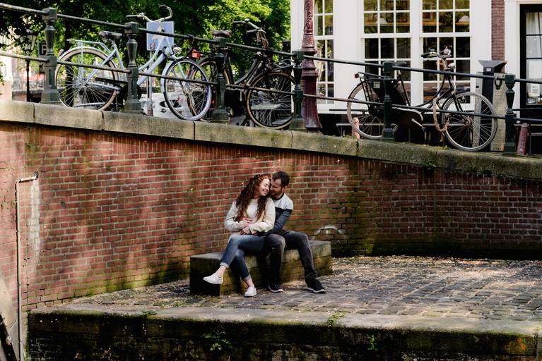 Couple sitting together by a quiet Amsterdam canal wall, sharing a warm moment before their engagement.