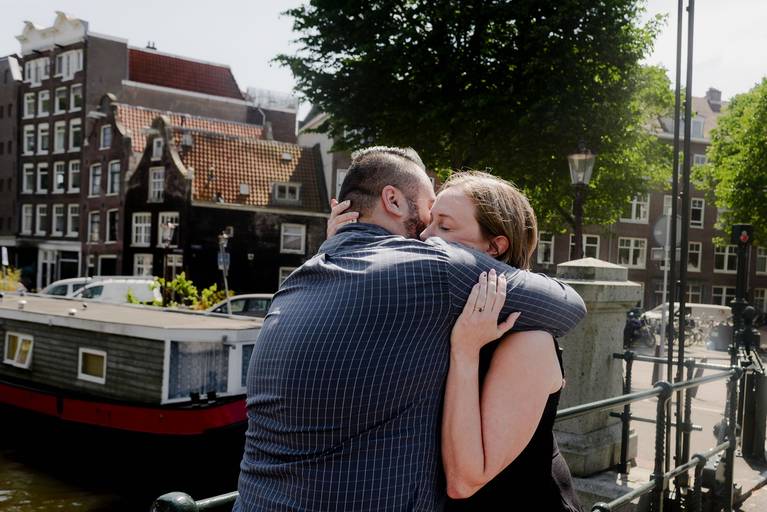 Emotional embrace on an Amsterdam bridge as the couple celebrates their engagement with canal houses behind them.