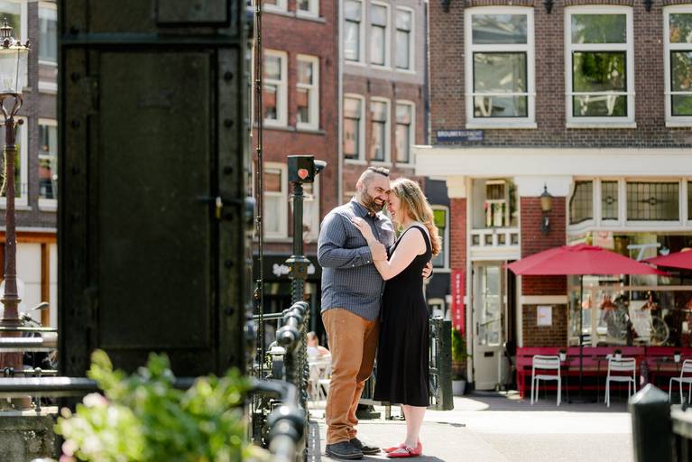 Engaged couple sharing a quiet moment on a sunny Amsterdam bridge with classic canal houses in the background.