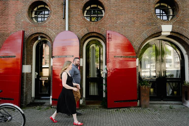 Couple walking past historic Dutch architecture with bright red shutters during an engagement photo session in Amsterdam.